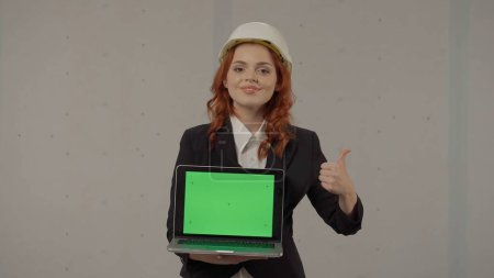 Téléchargez les photos : Une femme architecte tient un écran d'ordinateur portable tourné vers l'avant, donnant un pouce vers le haut. Portrait d'une femme d'affaires vêtue d'un casque et avec un ordinateur portable dans les mains sur un fond gris dans le studio. Publicité - en image libre de droit