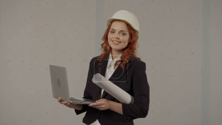 Foto de Una arquitecta trabaja en una laptop, estudia planos, imprime informes. Mujer de negocios con un casco protector, con dibujos bajo el brazo y un portátil sobre un fondo gris en el estudio - Imagen libre de derechos