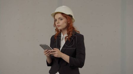 Téléchargez les photos : Une femme architecte utilise une tablette au travail. Femme d'affaires dans un casque de protection et avec une tablette sur un fond gris dans le studio - en image libre de droit