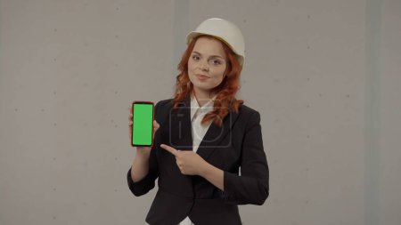 Téléchargez les photos : Une architecte féminine tient l'écran du smartphone tourné vers l'avant et pointant vers son écran vert. Portrait d'une femme d'affaires portant un casque sur fond gris en studio. Espace publicitaire - en image libre de droit