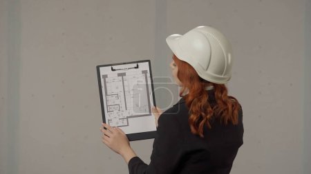 Photo for Back view of a woman architect with a apartment plan on a clipboard. A business woman studies the project plan and prepares for the renovation process. Woman in a protective helmet in the studio on a - Royalty Free Image