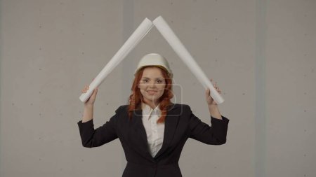 Foto de Una arquitecta eleva planos retorcidos en forma de casa sobre su cabeza. Mujer de negocios con traje y casco de construcción en el estudio sobre fondo gris - Imagen libre de derechos