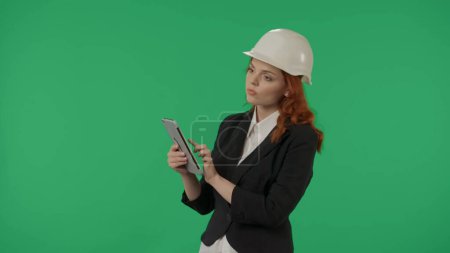 Foto de Una arquitecta realiza una inspección del objeto y toma notas en la tablilla. Mujer de negocios en un casco protector y con una tableta en una pantalla verde en el estudio - Imagen libre de derechos