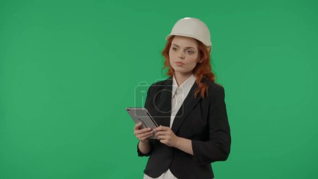 Téléchargez les photos : Une femme architecte dans un casque de protection et avec une tablette sur un fond vert écran dans le studio. Espace publicitaire, maquette d'espace de travail - en image libre de droit