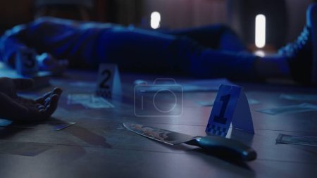 Foto de Escena del crimen con un cadáver y un cuchillo ensangrentado en el suelo en un apartamento oscuro de cerca. Un cuchillo ensangrentado y billetes de dólar están esparcidos por el suelo y marcados como evidencia, iluminados por la luz azul - Imagen libre de derechos
