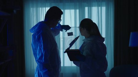 Photo for Male and female forensic investigator use a tablet to list evidence found at a crime scene in a dark apartment. A team of forensic examiners in protective lab suits and goggles, illuminated by blue - Royalty Free Image