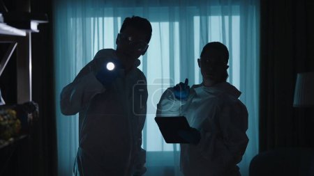 Photo for Forensic experts man and woman use a flashlight tablet in work at a crime scene in a dark apartment. A team of forensic examiners in protective lab suits and goggles, illuminated by blue light from - Royalty Free Image