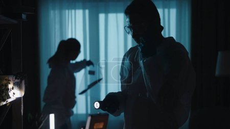 Photo for A team of forensic specialists work at the crime scene, in a dark apartment lit by blue light from police sirens. A man with a flashlight speaks on his smartphone, while a woman examines the murder - Royalty Free Image