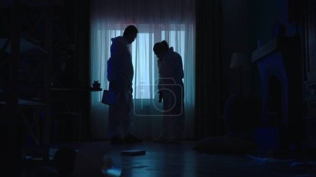 Photo for A team of forensic experts in protective suits and glasses use a flashlight as they scan the crime scene for clues. Silhouettes of professionals, a man and a woman in a dark apartment lit by blue - Royalty Free Image
