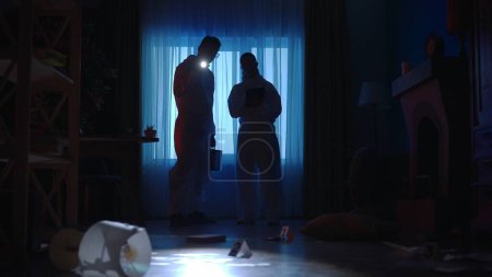 Photo for Male and female forensic investigator use a tablet and flashlight to list evidence found at a crime scene in a dark apartment. A team of forensic examiners in protective lab suits and goggles - Royalty Free Image