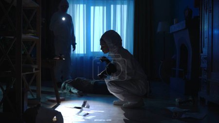 Photo for A team of forensic scientists collect evidence at the crime scene, in a dark apartment lit by blue light. A man inspects a crime scene using a flashlight. A woman photographs evidence with a camera - Royalty Free Image