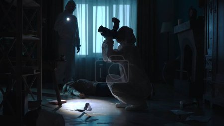 Foto de Un equipo de científicos forenses recoge pruebas en la escena del crimen, en un apartamento oscuro iluminado por la luz azul. Un hombre inspecciona una escena del crimen usando una linterna. Una mujer fotografía pruebas con una cámara - Imagen libre de derechos