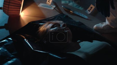 Foto de La mujer asesinada yace en una bolsa de cadáveres en el suelo, junto a la evidencia dispersa. El cadáver de una mujer en un apartamento oscuro en el suelo - Imagen libre de derechos