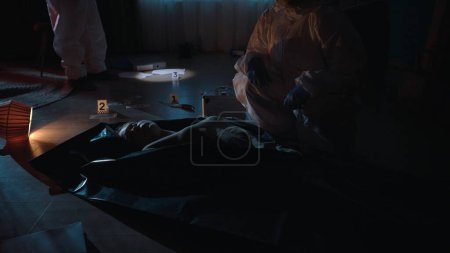 Foto de Un equipo de expertos forenses trabaja en una escena del crimen en un apartamento oscuro. Uno de los especialistas examina el cuerpo de la víctima en una bolsa de cadáveres, el otro examina la evidencia en la habitación con un - Imagen libre de derechos