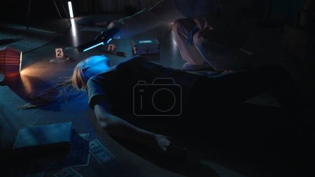 Téléchargez les photos : L'expert médico-légal se penche sur le corps des femmes et utilise une lampe ultraviolette pour rechercher des traces et des échantillons d'ADN. Un criminologue en combinaison de protection et avec une valise de travail travaille sur une scène de crime - en image libre de droit