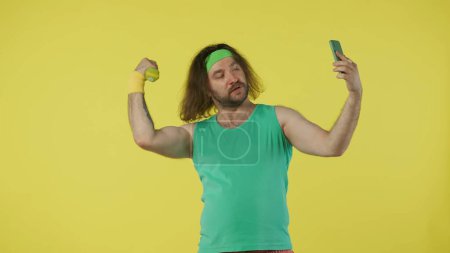 Téléchargez les photos : Homme en tenue de sport prenant selfie avec haltères. Portrait du modèle masculin en débardeur vert et bandeau. Concept fitness et bien-être. Isolé sur fond jaune. - en image libre de droit