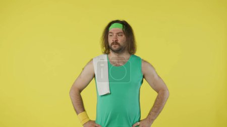 Téléchargez les photos : Homme en tenue de sport posant avec une serviette blanche. Portrait du modèle masculin en débardeur vert et bandeau. Concept fitness et bien-être. Isolé sur fond jaune. - en image libre de droit