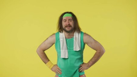 Photo for Man in sportswear standing smiling, white towel over neck. Portrait of male model in green tank top and headband. Fitness and wellness concept. Isolated on yellow background. - Royalty Free Image