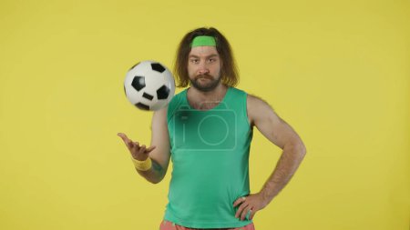 Téléchargez les photos : L'homme en tenue de sport tenant le ballon de football et le vomit dans la main. Portrait du modèle masculin en débardeur vert et bandeau. Concept fitness et bien-être. Isolé sur fond jaune. - en image libre de droit