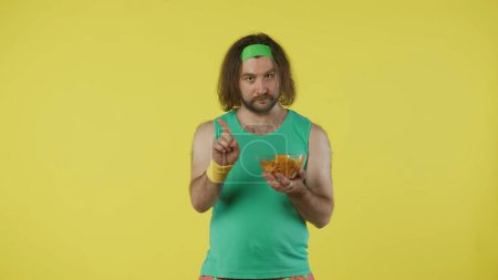 Photo for Man in sportswear holding a bowl of potato chips and shows negative sign. Portrait of male model in green tank top and headband. Fitness and wellness concept. Isolated on yellow background. - Royalty Free Image