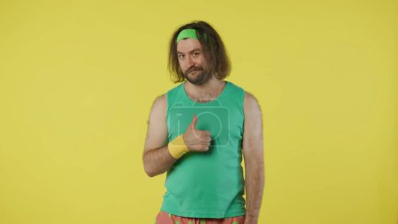 Photo for Man in sportswear looking at the camera, winking and showing thumbs up sign. Portrait of male model in green tank top and headband. Fitness and wellness concept. Isolated on yellow background. - Royalty Free Image