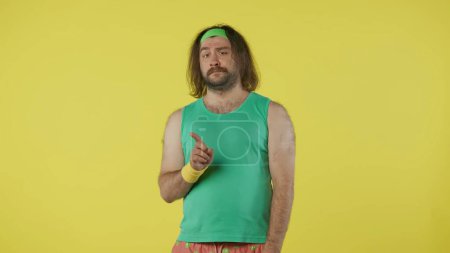 Photo for Man in sportswear looking at the camera, waving finger in negative way. Portrait of male model in green tank top and headband. Fitness and wellness concept. Isolated on yellow background. - Royalty Free Image