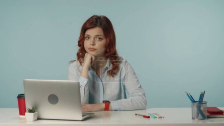Photo for Medium isolated shot of a dissatisfied and confused young woman sitting at the desk with laptop and working supplies, looking straight at the camera. She is leaning on her hand. Creative content. - Royalty Free Image