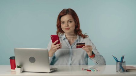 Photo for Medium isolated shot of a satisfied, happy and relaxed young woman, sitting at the desk, holding a phone and a creadit card. Concept of online shopping. Creative content or advertisement. - Royalty Free Image