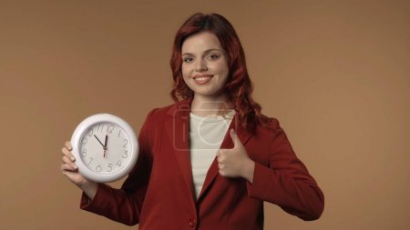 Photo for Medium isolated shot of a satisfied, happy and active young woman giving a thumbs up with a clock in her hand, smiling. Business content or advertisement. - Royalty Free Image