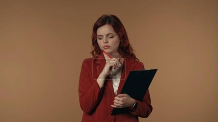 Photo for Medium isolated shot of a young woman pensively looking away and trying to come up with an idea, while holding a planchette, folder in one hand and a pecil next to her chin. Creative content or - Royalty Free Image
