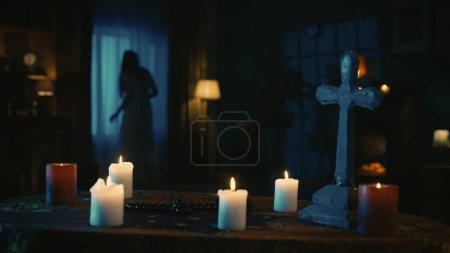 Téléchargez les photos : Prise de vue d'une table avec des outils magiques : bougies, croix et perles. Sur le fond, il y a une silhouette féminine floue, fantôme se déplaçant bizarrement devant la fenêtre. Contenu créatif. - en image libre de droit