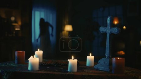 Photo for Shot capturing a table with magical tools on it: candles, cross and beads. On the background there is a blurry female silhouette, ghost moving weirdly in front of the window. Creative content. - Royalty Free Image