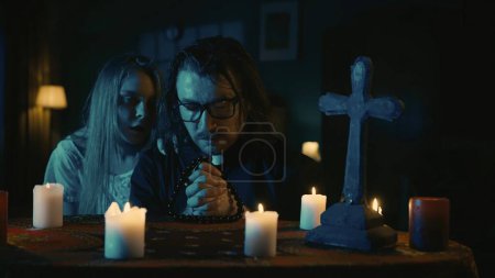 Photo for Shot capturing a table with magical tools on it. A priest is sitting at the table with beads in his hands, praying with his eyes closed. Female entity, ghost stands from behind. Creative content. - Royalty Free Image