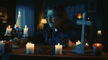 Photo for Shot capturing a priest sitting at the table, looking down with a candlestick and beads in his hands, prepared to perform a rite, ritual. Creative content, halloween themed. - Royalty Free Image