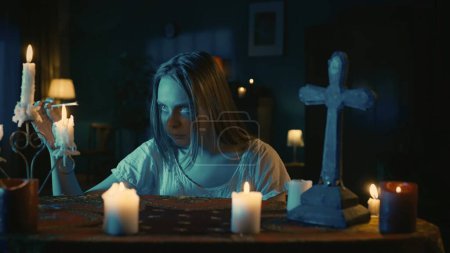 Photo for Shot capturing a young woman sitting at the table, surrounded by candles, crosses and beads, holding a lit match in her hand, prepared to perform a rite, ritual. Creative content, halloween themed. - Royalty Free Image