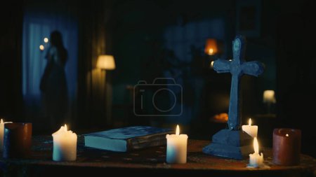 Téléchargez les photos : Prise de vue d'une table avec des outils magiques : bougies, croix et perles. Une jeune femme, entité avec un chandelier, se tient près de la fenêtre. Place pour les rites et rituels occultes. Contenu créatif - en image libre de droit