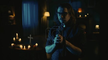 Photo for Shot capturing a priest standing in the dark room with beads and a cross in his hands, praying with his eyes closed. On the background there is a table with magical tools on it. Creative content. - Royalty Free Image