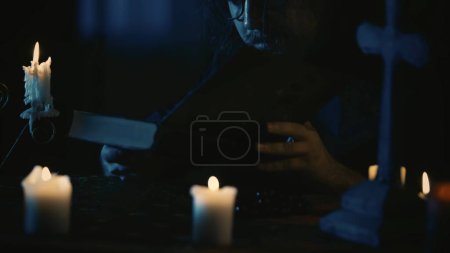 Photo for Shot capturing a priest sitting at the table, surrounded by candles, crosses and beads, holding a book in hands, reading it carefully. The man is performing a rite, ritual. Creative content, halloween - Royalty Free Image