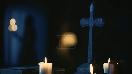Téléchargez les photos : Prise de vue d'une table avec des outils magiques : bougies, croix et perles. Une jeune femme, entité avec un chandelier, se tient près de la fenêtre. Place pour les rites et rituels occultes. Contenu créatif - en image libre de droit