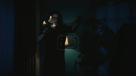 Photo for Shot capturing a priest holding a candlestick and a cross in hands, scaking them to scare something he sees off the frame. The man is frightened. Creative content, halloween themed. - Royalty Free Image