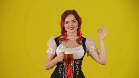 Foto de Fondo amarillo medio plano aislado de una joven alemana, camarera, vestida con un traje tradicional, sosteniendo un vaso de cerveza, mostrando el signo OK y sonriendo. Festival de octubre, festival de la cerveza, publicidad - Imagen libre de derechos
