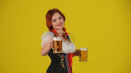 Foto de Fondo amarillo medio plano aislado de una joven alemana, camarera, vestida con un traje tradicional, sosteniendo dos vasos de cerveza, ofreciendo uno y sonriendo. Festival de octubre, festival, publicidad. - Imagen libre de derechos