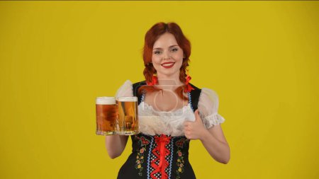 Foto de Fondo amarillo medio plano aislado de una joven alemana, camarera, con un traje tradicional, sosteniendo un vaso de cerveza, cerveza, dando un pulgar hacia arriba y sonriendo. Octoberfest, festival de la cerveza. - Imagen libre de derechos