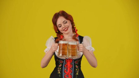 Foto de Fondo amarillo medio plano aislado de una joven alemana, camarera, vestida con un traje tradicional, haciendo clic en dos vasos de cerveza, animando y sonriendo. Festival de octubre, festival de la cerveza, publicidad. - Imagen libre de derechos