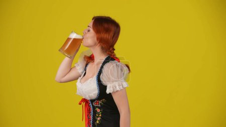 Photo for Medium yellow background isolated side-view shot of a young German woman, waitress, wearing a traditional costume, taking a sip, gulp of beer with pleasure. Octoberfest, beer festival, advertisement. - Royalty Free Image