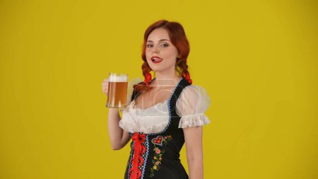 Foto de Fondo amarillo medio plano aislado de una joven alemana, camarera, vestida con un traje tradicional, sosteniendo un vaso de cerveza, lager y lamiéndose los labios. Festival de octubre, festival de la cerveza, publicidad. - Imagen libre de derechos