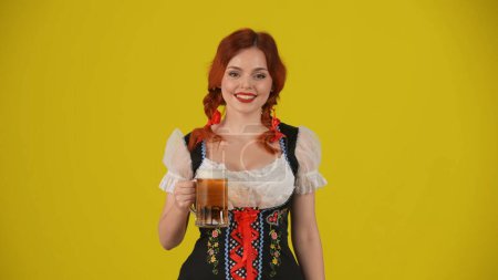 Téléchargez les photos : Fond jaune moyen plan isolé d'une jeune Allemande, serveuse, vêtue d'un costume traditionnel, tenant un verre de bière, de bière et souriante. Octoberfest, festival de la bière, publicité. - en image libre de droit