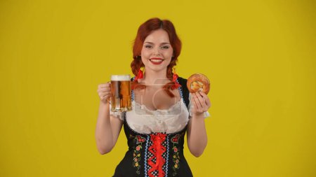 Foto de Fondo amarillo medio plano aislado de una joven alemana, camarera, vestida con un traje tradicional, sosteniendo un pretzel y un vaso de cerveza y sonriendo. Festival de octubre, festival de la cerveza, publicidad. - Imagen libre de derechos