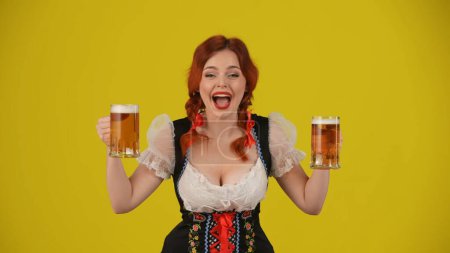Foto de Fondo amarillo medio plano aislado de una joven alemana, camarera, vestida con un traje tradicional, sosteniendo dos vasos de cerveza, mostrando wow y emoción. Octoberfest, publicidad. - Imagen libre de derechos