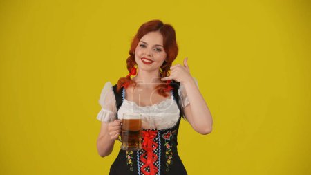 Photo for Medium yellow background isolated shot of a young German woman, waitress wearing a traditional costume, holding a glass of beer, looking at the camera and showing call me gesture. Advertisement. - Royalty Free Image
